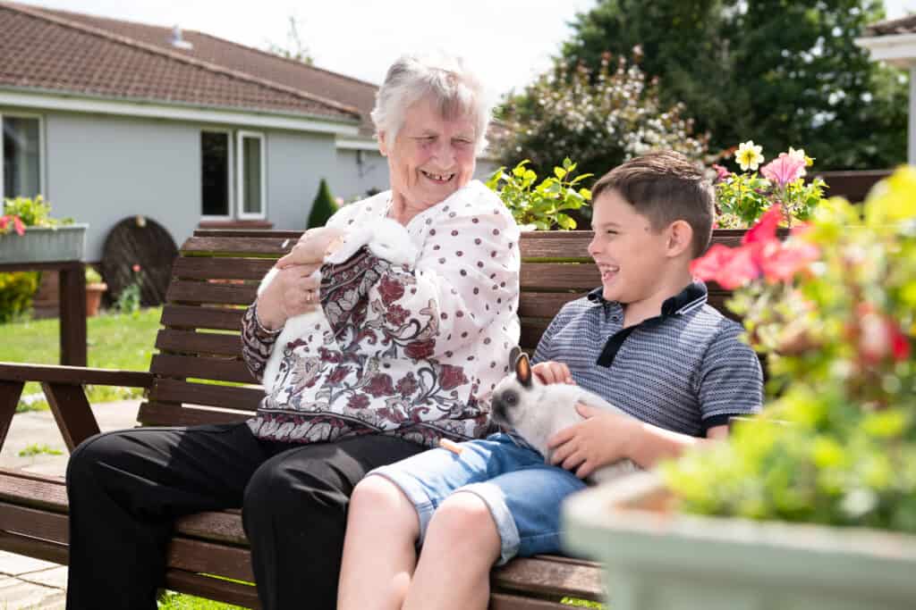 Boy sits on bench in care home garden with older woman and they stroke a bunny rabbit