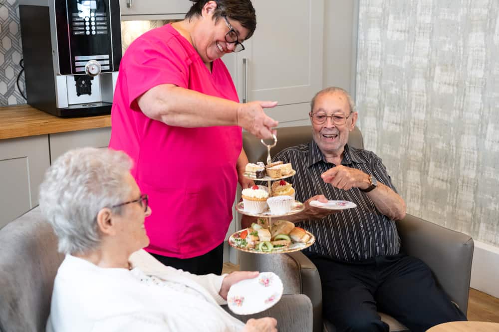 A carer in pink uniform serves cakes to older people in armchairs